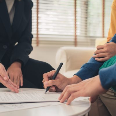 Happy young Asian couple and realtor agent. Cheerful young man signing some documents while sitting at desk together with his wife. Buying new house real estate. Signing good condition contract.